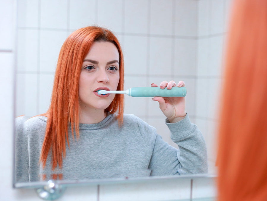 Chica de cabello pelirrojo se cepilla los dientes delante del espejo.